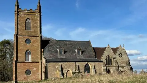 Bridget Weaving A two-storey church with three story tower and a pitched slate roof with dormer windows. It is in a field, with tall grass. The picture is taken on a sunny day, with light clouds and blue sky.