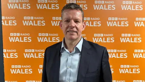 Rhun ap Iorwerth pictured standing in front of a large orange board with BBC Radio Wales logos all over it. He is smiling and wearing a dark suit jacket and light blue shirt. 