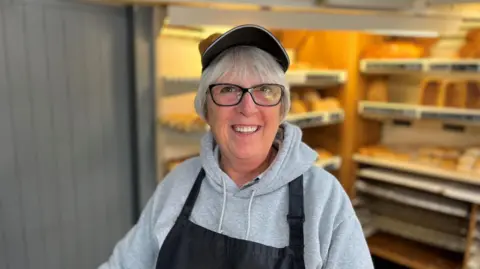 A woman with grey hair, a cap, a light grey top and a black apron standing in front of shelves with bread on them