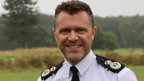 A man with dark hair and a smile on his face in a white shirt with police badges on his shoulders, standing in a field with trees behind him