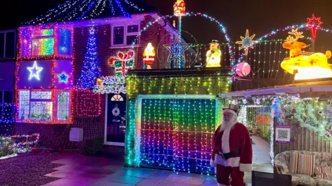 Danny Fullbrook/BBC A wider shot of Mr O'Toole's 1950s-style semi-detached house covered in Christmas lights. Mr O'Toole is on the left dressed as Santa, standing at the entrance of a grotto.