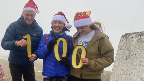 Laura Nuttall's parents and sister - all in Santa hats - hold up the number 100 on top of Pendle Hill. They are all smiling. The rocks beside them and the sky are both grey.