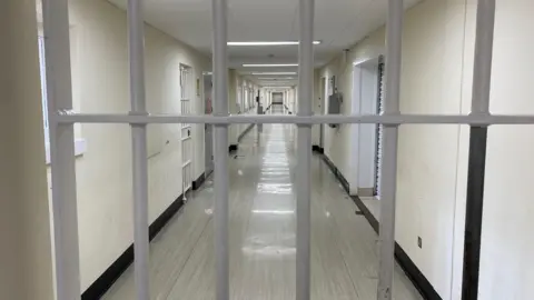 A barred door blocks the photographer's way. There is a grey floor and white walls lining a corridor heading straight into the back of the shot with a barred door to the left and white shutters to the right.