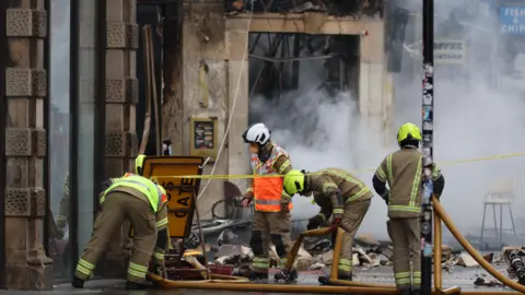 PA Media A team of firefighters on the pavement in front of a collapsed building. Rubble lies on the ground and there is smoke in the background.