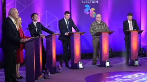 Five men and one woman behind lecterns on stage at the debate in Wrexham with a BBC election logo in the purple background. 