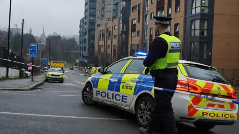 A police incident scene on a city street, with emergency services and a road closure in place. Modern apartment buildings line the right side of the road. These buildings have a mix of brick and grey panel exteriors, multiple floors, and rows of windows and balconies. In the foreground, a police car is parked diagonally across the road, partially blocking traffic. The vehicle is marked with high-visibility yellow and blue police livery, and the word “POLICE” is clearly visible on the side and rear. The car’s blue emergency lights are flashing.