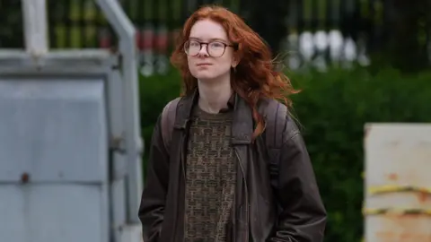 A woman with ginger hair and glasses wearing a brown jacket and brown jumper is walking, wearing a black rucksack.