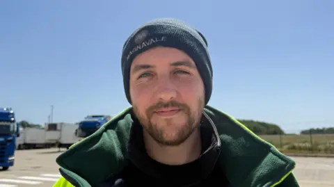 A man with a brown beard stands in a yard on a sunny day while wearing a green woolly hat and a yellow and green coat. HGVs can be seen behind him and the sky is blue.