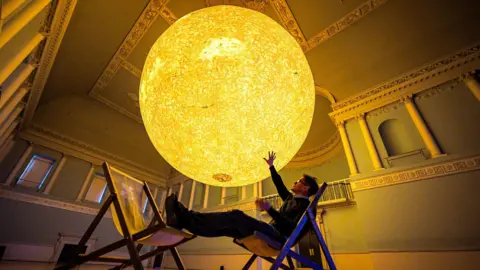 PA Media The artist Luke Jerram sits in a deckchair with his feet on another deckchair underneath a large glowing replica of the sun. The photograph is taken inside the Assembly Rooms in Bath. The artwork is called Helios