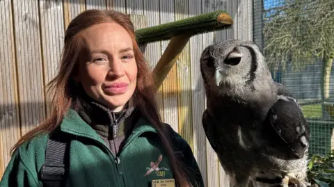 George King/BBC Emily Quantrill wearing a green fleece and holding a large owl inside an enclosure. She is looking into the camera.