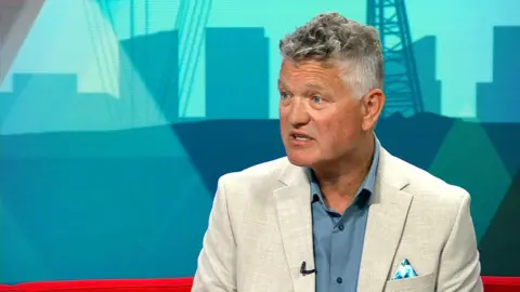 Mike Starkie sits in front of the teal set of BBC Politics North wearing a beige suit, dusty blue shirt and a vibrant patterned pocket handkerchief. 