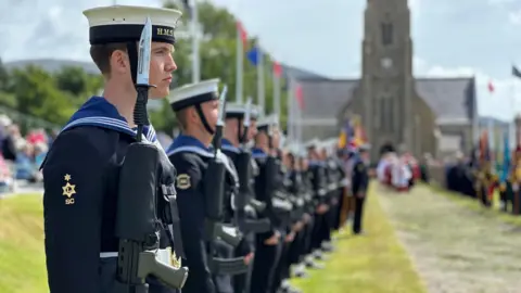 Members of the guard of honour lining the ceremonial walkway wearing blue Royal Navy uniforms and white hats and holding guns with bayonets. Members of the clergy in white and red robes are emerging from the Royal Chapel in the distance.