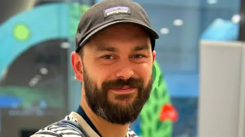 Sew Positive Adam is smiling at the camera while inside. His face is cast in a warm orange glow from the lighting. He has a blue and white stripy top on and a grey Patagonia cap. He has brown eyes and a dark brown beard. 