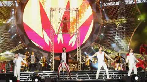 Take That, with four members, are pictured on stage during a live performance. All four members of the band appear to be singing into microphones on stands. Behind them there is a large circus-themed set, including a tall structure with trapeze artists at the top. 