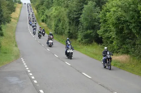 On a hilly road, many motorbikes ride in succession, with grass and trees either side of the road.
