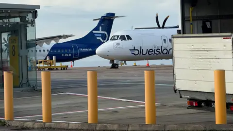 BBC Two Blue Island planes on the tarmac at an airport. Large yellow bollards are in the foreground of the photo. The back of a lorry parked on the runway can be seen just behind the bollards.