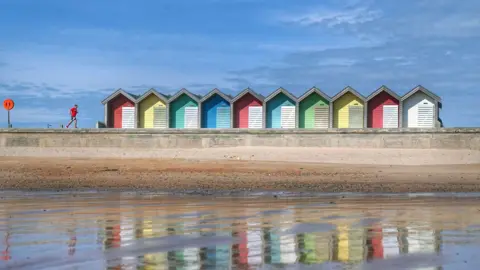 Blyth beach at low tide, looking back to the beach from the water. Colourful huts line the beach and a man with a red top runs along the promenade. The huts are reflected in the water and the sky is very blue. 