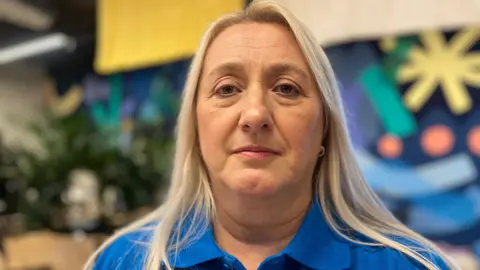 A blonde hair woman looks at the camera. She wears a blue polo shirt and a gold earing, in her left ear. Over her right shoulder is a green bush, which sits in front of a multi-coloured wall.