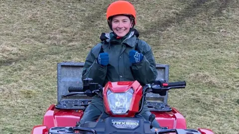 Rosie Ashworth A young woman wearing waterproof gear and an orange helmet sits on a red quad bike. She smiles and holds two thumbs up. In the background, a grassy hill can be seen.