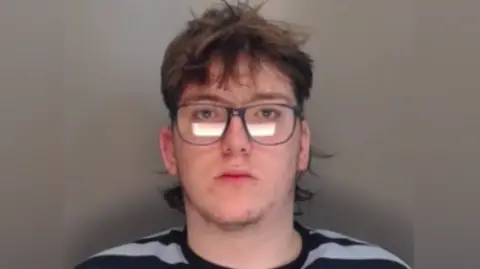 Joseph Thompson has a brunette mullet hair cut and wears large navy-rimmed glasses. He has a spotty appearance and has small amounts of facial hair. He is wearing a black and white striped t-shirt.