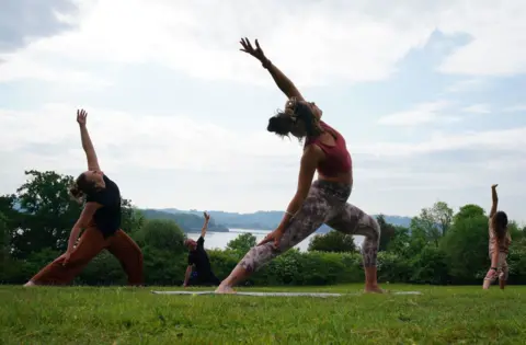 Three woman stretch in yoga poses on a green space next to Chew Valley Lake