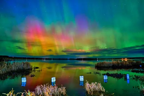 John Cuthbert The aurora, which has green, pink, yellow and blue colours, shining over a loch with lanterns in the foreground.