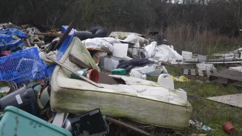 A large pile of fly‑tipped rubbish in a grassy area, including an old stained mattress, plastic crates, broken furniture and assorted household waste.