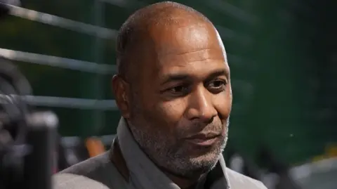 Les Ferdinand in a grey coat. He stands outside the pitches in Islington