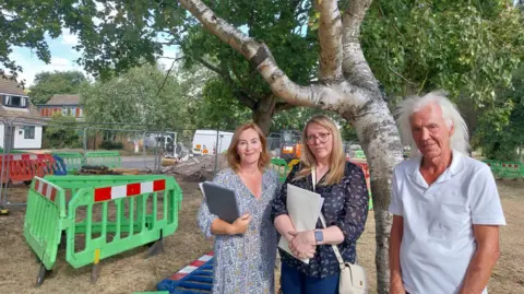Nadia Lincoln/LDRS Three people are standing in front of a large tree. Two women are dressed in different blue outfits and holding paperwork. A man is standing beside them in a white T-shirt. There are plastic barriers and metal railings in the area to one side of the group.