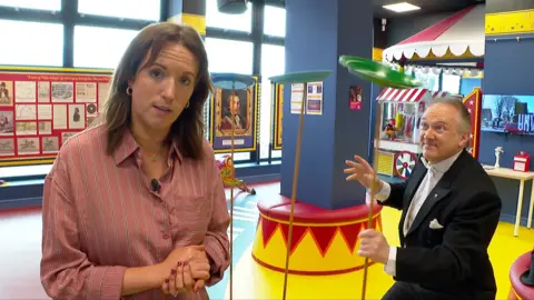 A woman in a pink top and long brown hair next to a circus performer in black tuxedo spinning plates