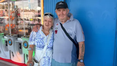 An older couple with their arms around one another. They're both holding a crutch in front of an amusement arcade. Both are in a pale blue colour. Pauline is in a floral dress with sunglasses on her head, her husband is wearing a cap and smiling.