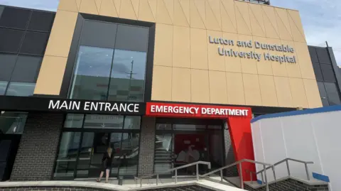 Bedfordshire Hospitals NHS FT The outside of The Luton and Dunstable Hospital, showing one person walking into an entrance. The building is black brick, with a large beige front. White boarding is to the right and metal railings. 
