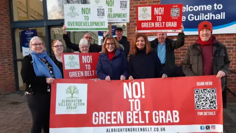 LDRS A group of people standing outside the Shropshire Council building that has a blue and white sign on the wall. They are smiling and celebrating, holding signs and boards that read "no to overdevelopment" and "no to green belt grab"