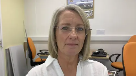 Jane Hewer is wearing rimless glasses and a white collared shirt. She has short blonde hair and is sitting in an office.