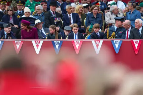PA Media (L-R) Veteran Bernard Morgan, the Princess of Wales, Princess Charlotte, Prince George, Prince Louis and the Prince of Wales sit on the front row of benches of a crowd outside Buckingham Palace with bunting saying '80' at the front.