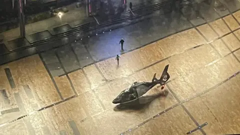 MICH2255 Two dark figures are walking away from the helicopter and towards a giant screen on the piazza.