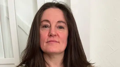 This image shows a woman with long brown hair and hazel eyes, she's sat infront of the a white wall.