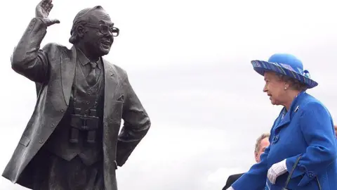 Getty Images Queen Elizabeth II, dressed in blue coat and hat, is standing near a statue of Eric Morecambe