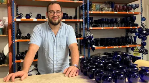 A man with a beard and glasses, wearing a sky blue shirt, stands proudly beside a table full of Bristol Blue Glass that he has made. The shelves behind him are stacked high with other samples of the same blue glass. 