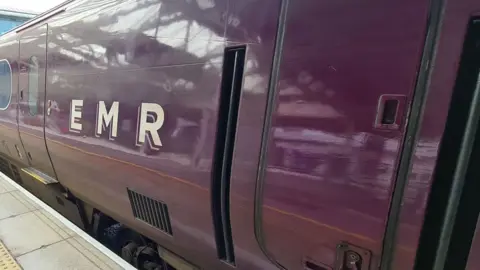 Oli Constable/BBC A close up of the side of an East Midlands carriage. It is branded in purple colour and has E M R written on its side in capital letters. It is sitting at a platform, which can just be seen on the left. 