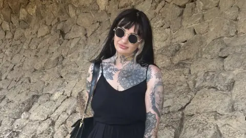 Daisy Farrar-Hayton Daisy Farrar-Hayton with long dark hair and sunglasses leaning against a stone wall