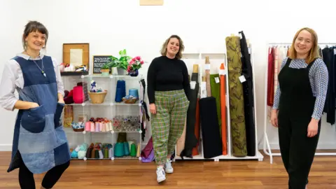 Photo + Flourish Three women stand in front of some sewing supplies.