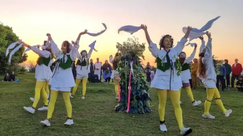 Annie Blick Morris dancers at Rodborough Common dancing an waving handkerchiefs with musicians and people watching with the sun rising in the background.