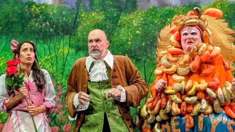 Belle, her father and panto dame Betty Bon Bon on stage at the Lawrence Batley Theatre with a castle meadow backdrop in the background. The panto dame is wearing a brightly coloured costume which looks like a tin of baked beans has been poured over them.