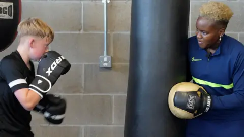Jamie Niblock/BBC A blonde haired boy wearing black boxing gloves prepares to punch the punchbag while Nicola stands next to it. 