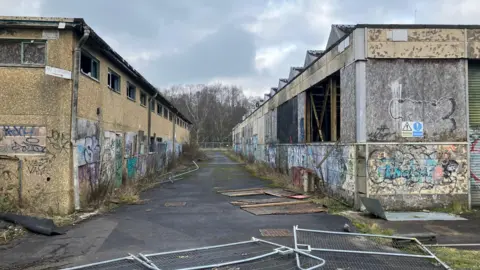 View of the derelict Saxonvale site in Frome, with buildings boarded up covered in graffiti. The buildings are lined up on either side and there are crash barriers lying on the floor