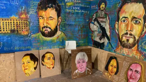 A board is painted blue with portraits of Ukrainian refugees and quotes, below are cardboard plaques with portraits of women painted on them.