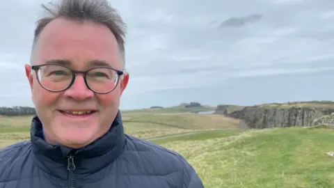 Tony Gates stands with a stretch of Hadrian's Wall rising on a crag behind him. He is a man in his 60s with glasses and short hair smiling and with a blue jacket zipped up to his neck.
