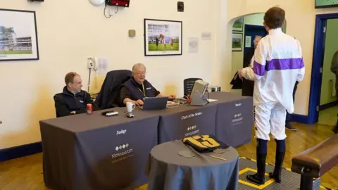 Qays Najm/BBC A jockey weighing room. A jockey wearing his uniform stands on a large scale. In front of him is a table where Lind sits in front of a laptop next to another man. 