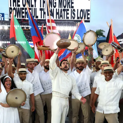 Getty Images Bad Bunny holding up a football with white writing, surrounded by musicians and dancers holding up instruments and flags
